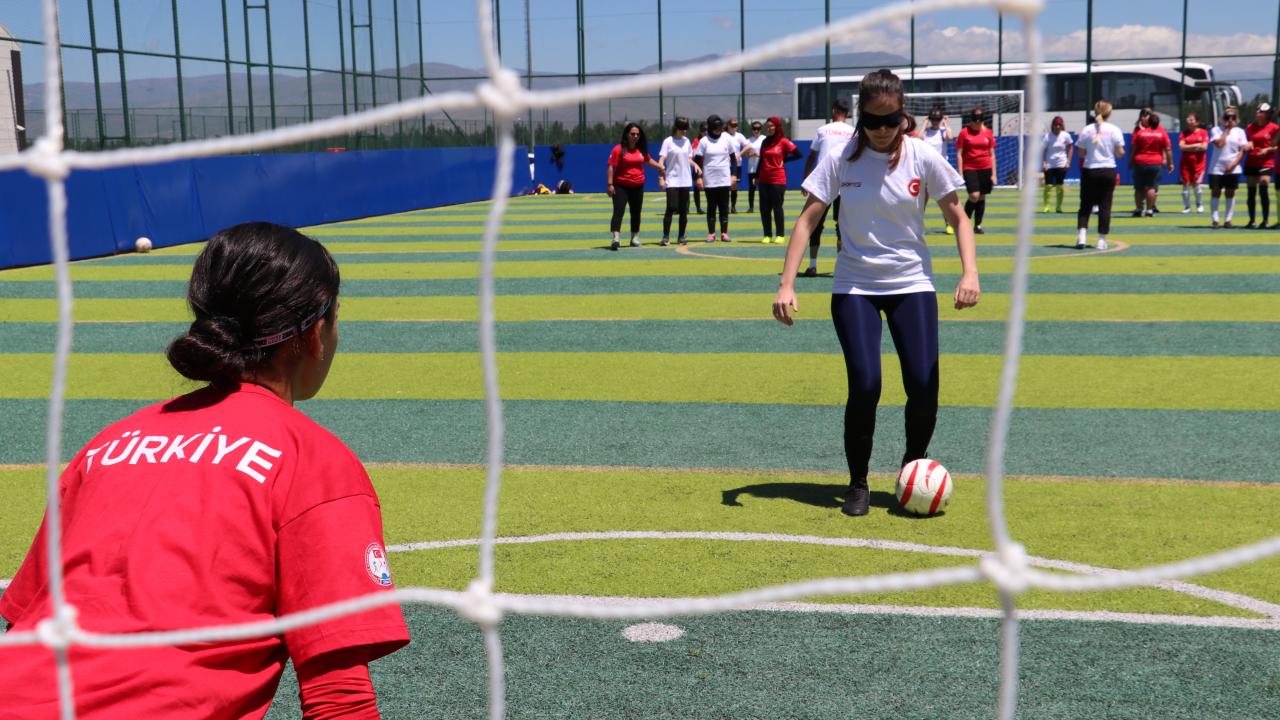 Fotoğrafta görme engelli kadın futbolcular var. Kaleci arkadan gözüküyor. Üzerinde Milli Takım forması var. Karşısında şut çekmek üzere olan bir başka futbolcu var. Gözleri bant ile kapalı.